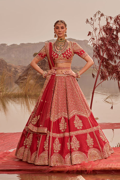 Woman in a red and gold traditional outfit standing on a platform by a lake with a scenic background.
