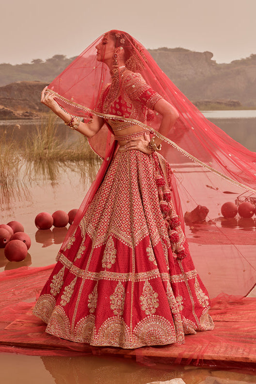 Woman in a red and gold traditional outfit standing on a platform over water with a scenic background.