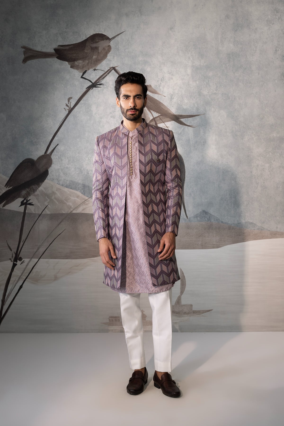 Man wearing a patterned kurta with white pants against a textured wall with birds.