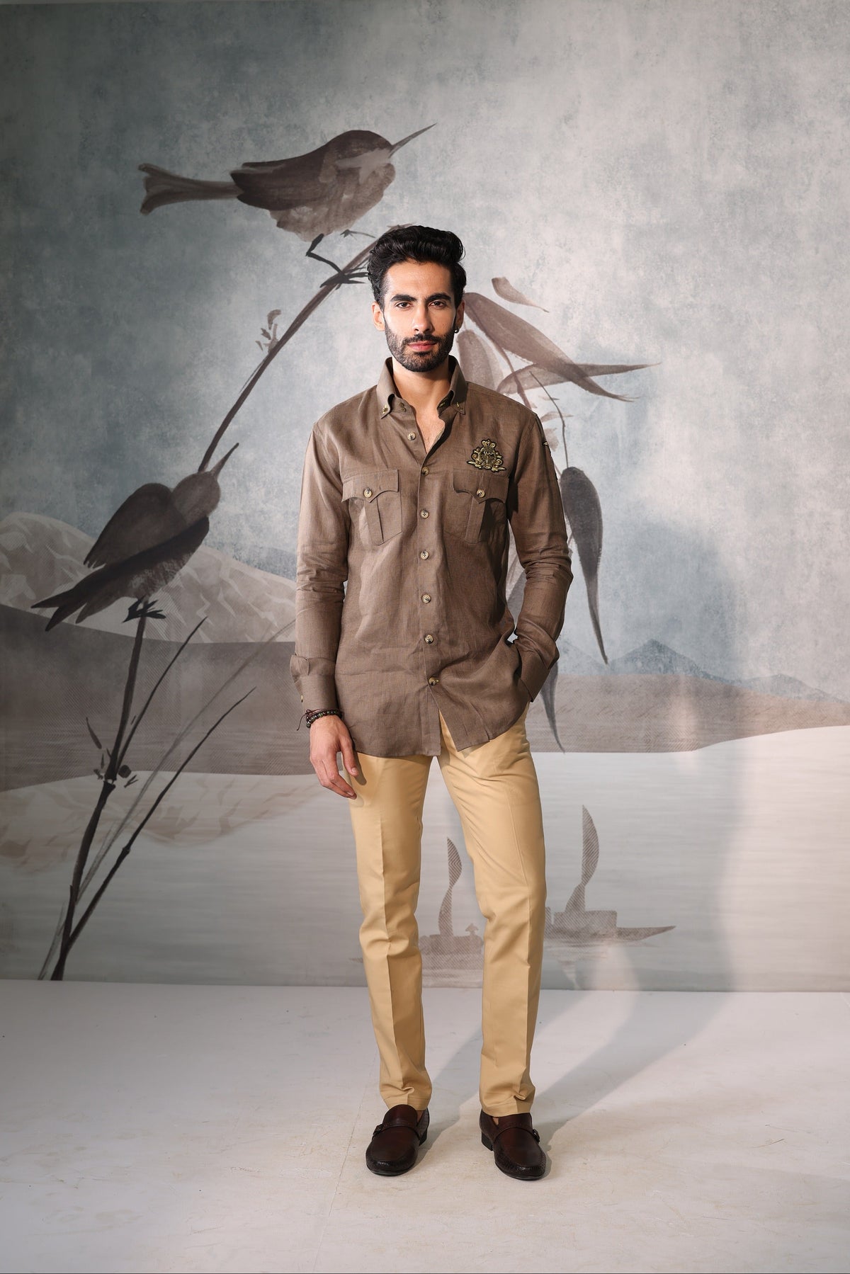 Man wearing a brown shirt and beige pants standing in front of a wall with bird artwork.