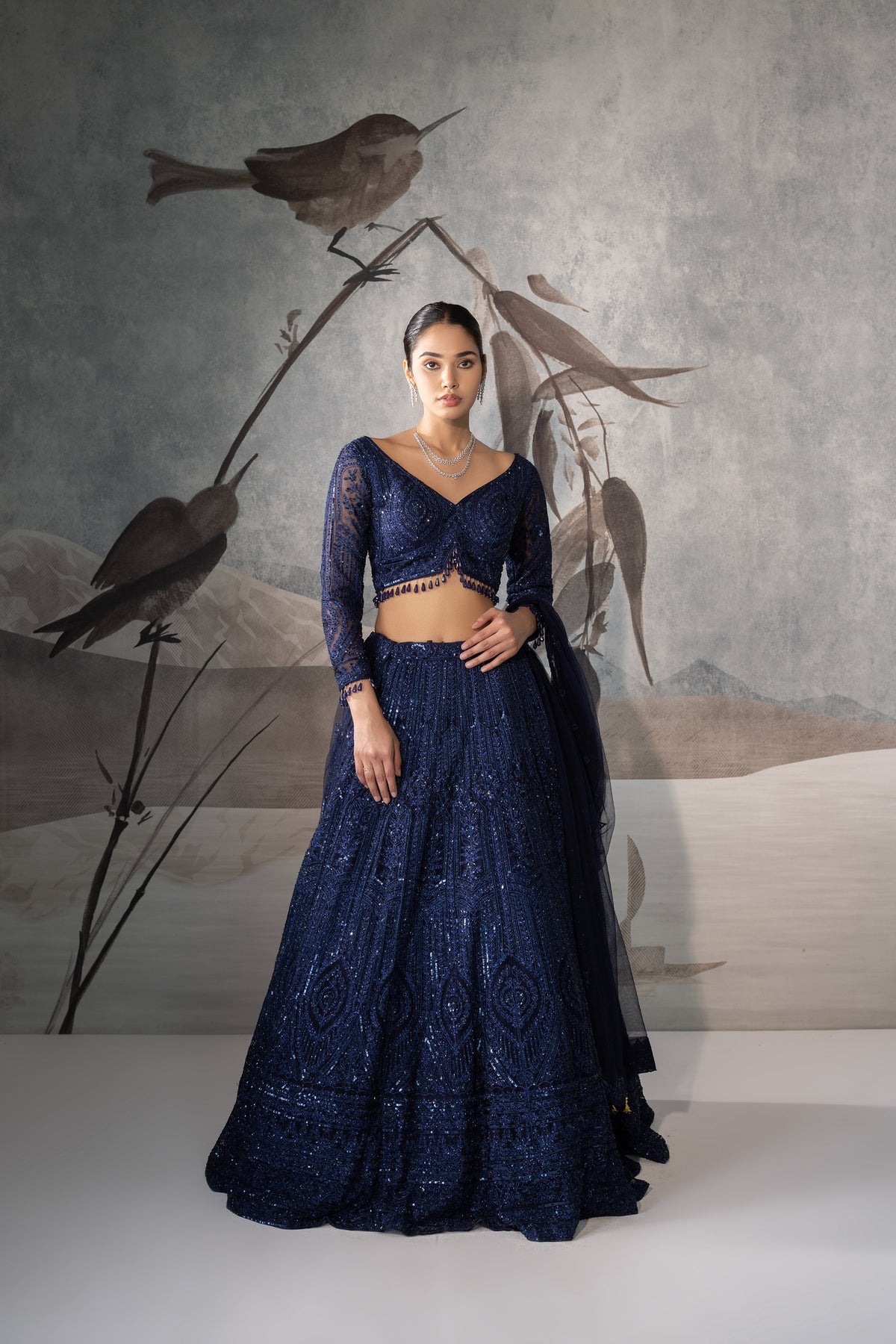 Woman in a dark blue embroidered outfit standing against a textured wall with bird decorations.