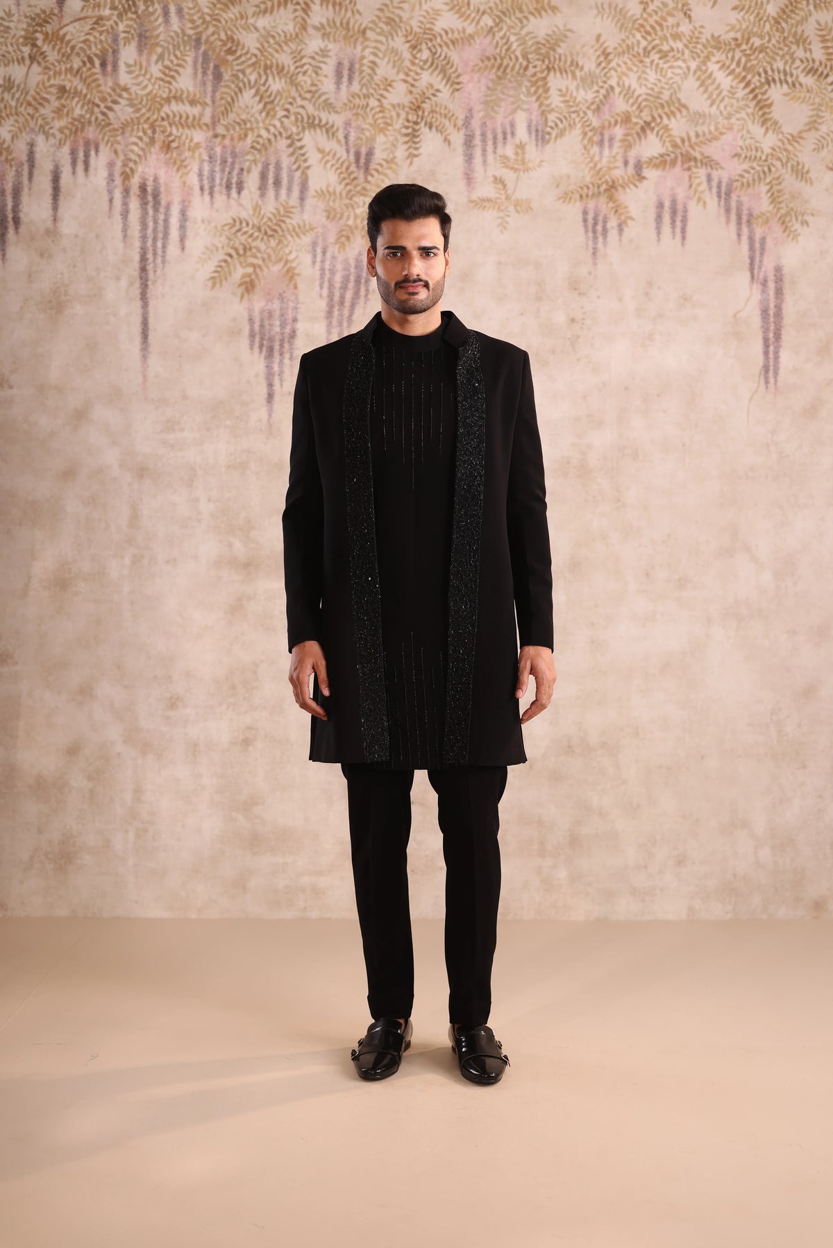 Man wearing a black coat and pants against a decorative wall.