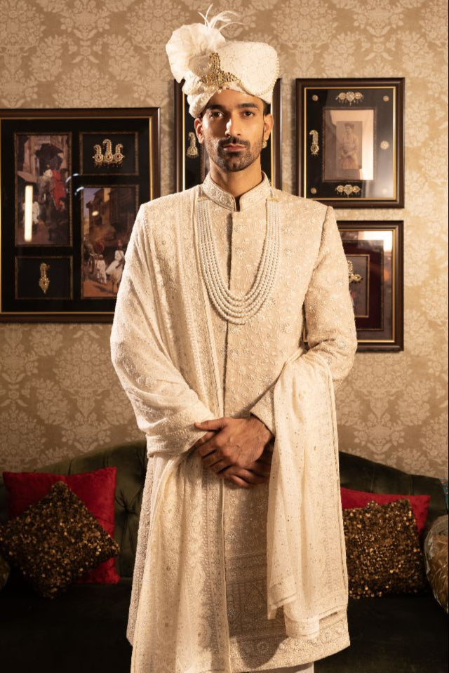 Man in traditional white sherwani with decorative turban in a decorated room.