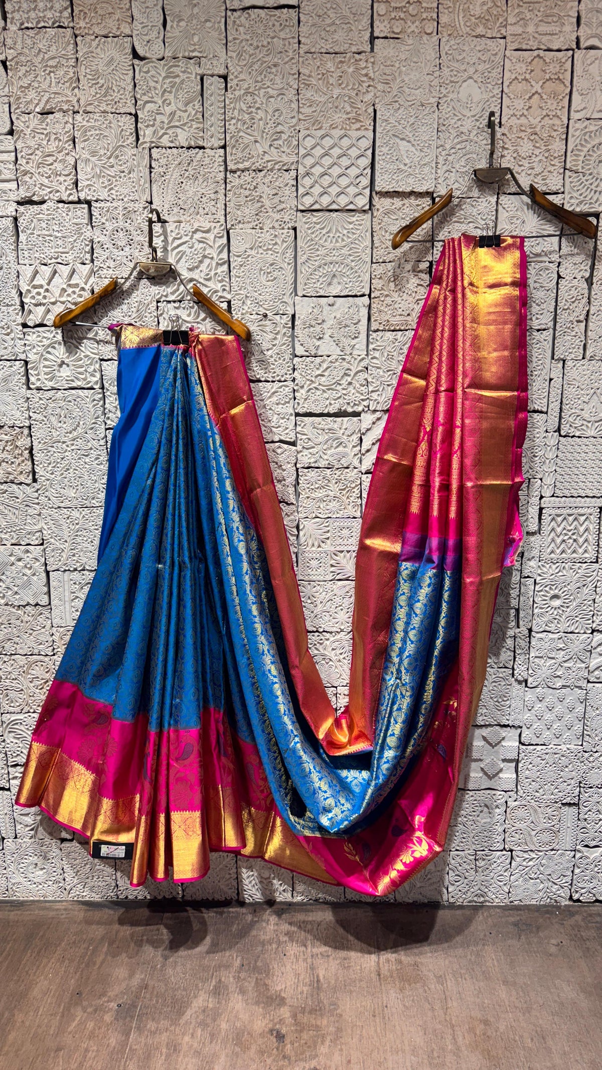 A royal blue and pink Kanjivaram silk saree with gold zari weaving, displayed on hangers against a white stone wall background.