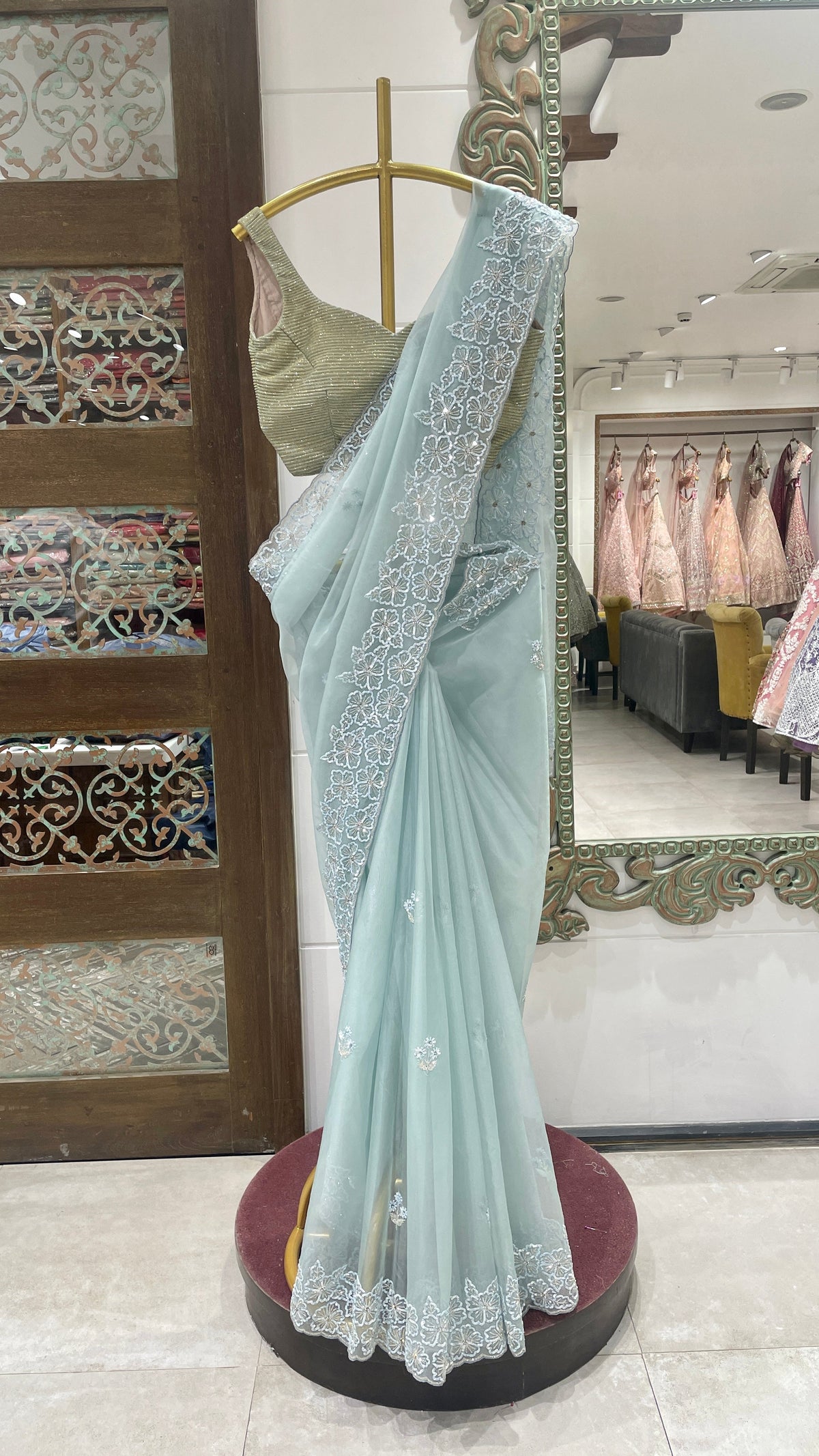A pastel blue organza saree with embellishments displayed on a mannequin, with the saree's end showing detailed floral pattern and embellishments.
