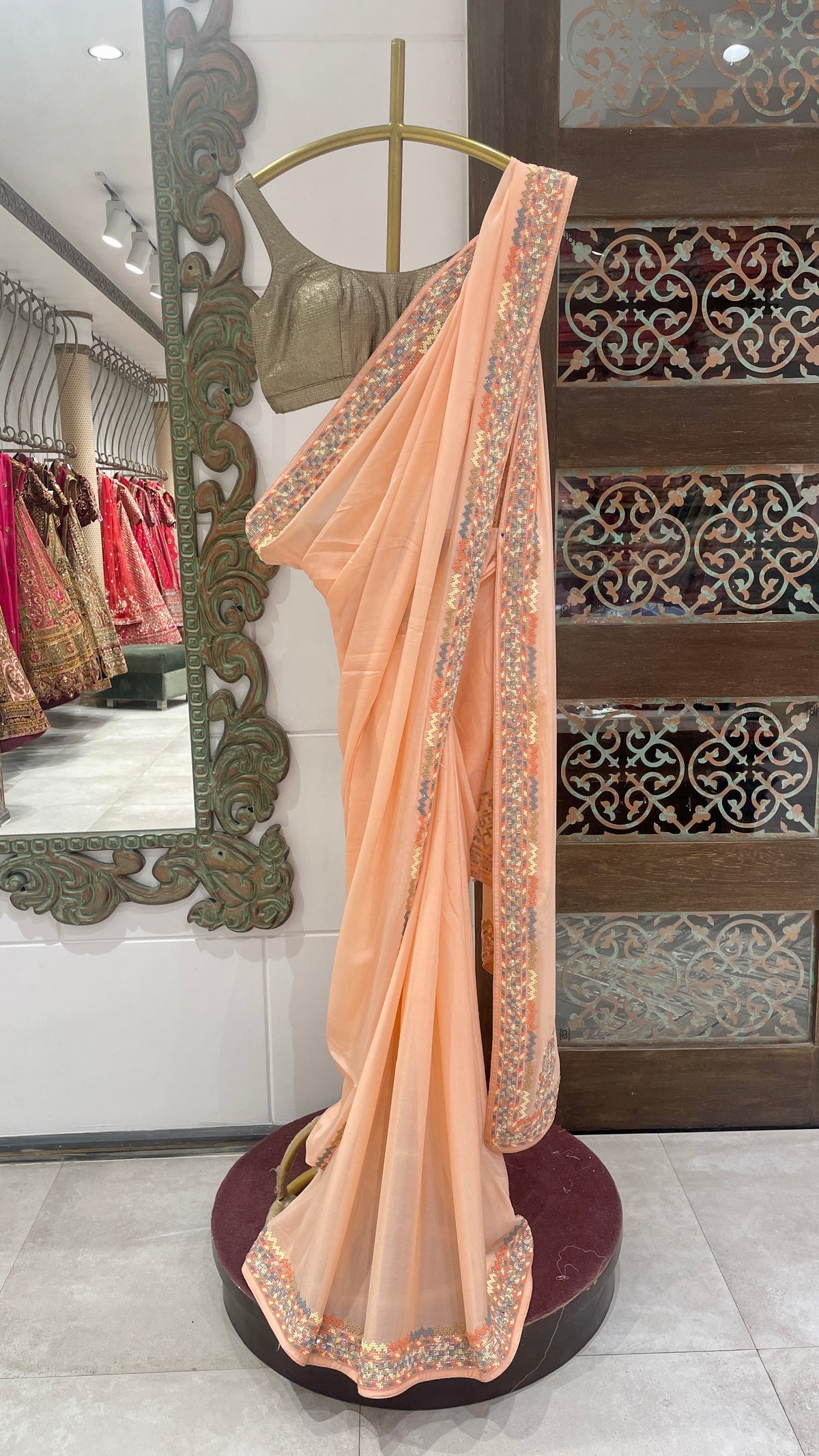A peach crepe saree with embellishments displayed on a mannequin in a store with intricate floral and geometric patterns on the fabric.