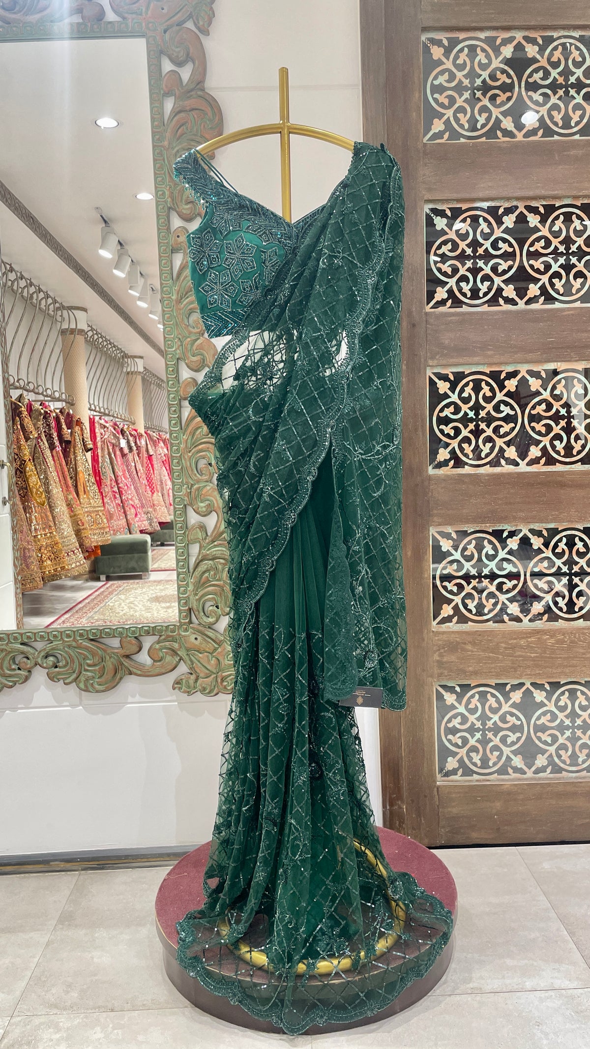 A green net saree with floral embellishments displayed on a mannequin in a store with traditional attire and decorative patterns in the background.