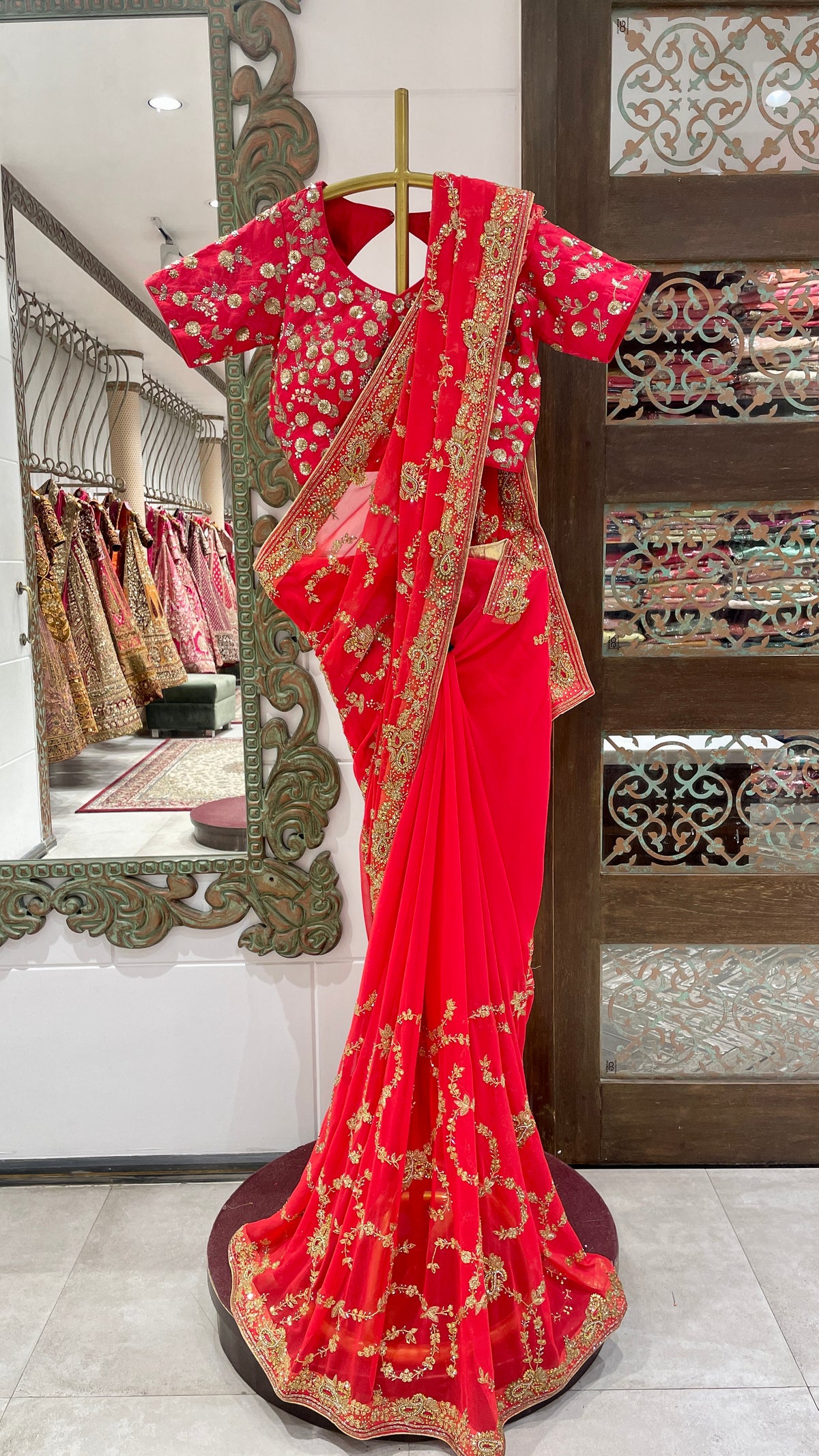 A red saree with gold embellishments draped over a mannequin, displayed in a store with an ornate background.