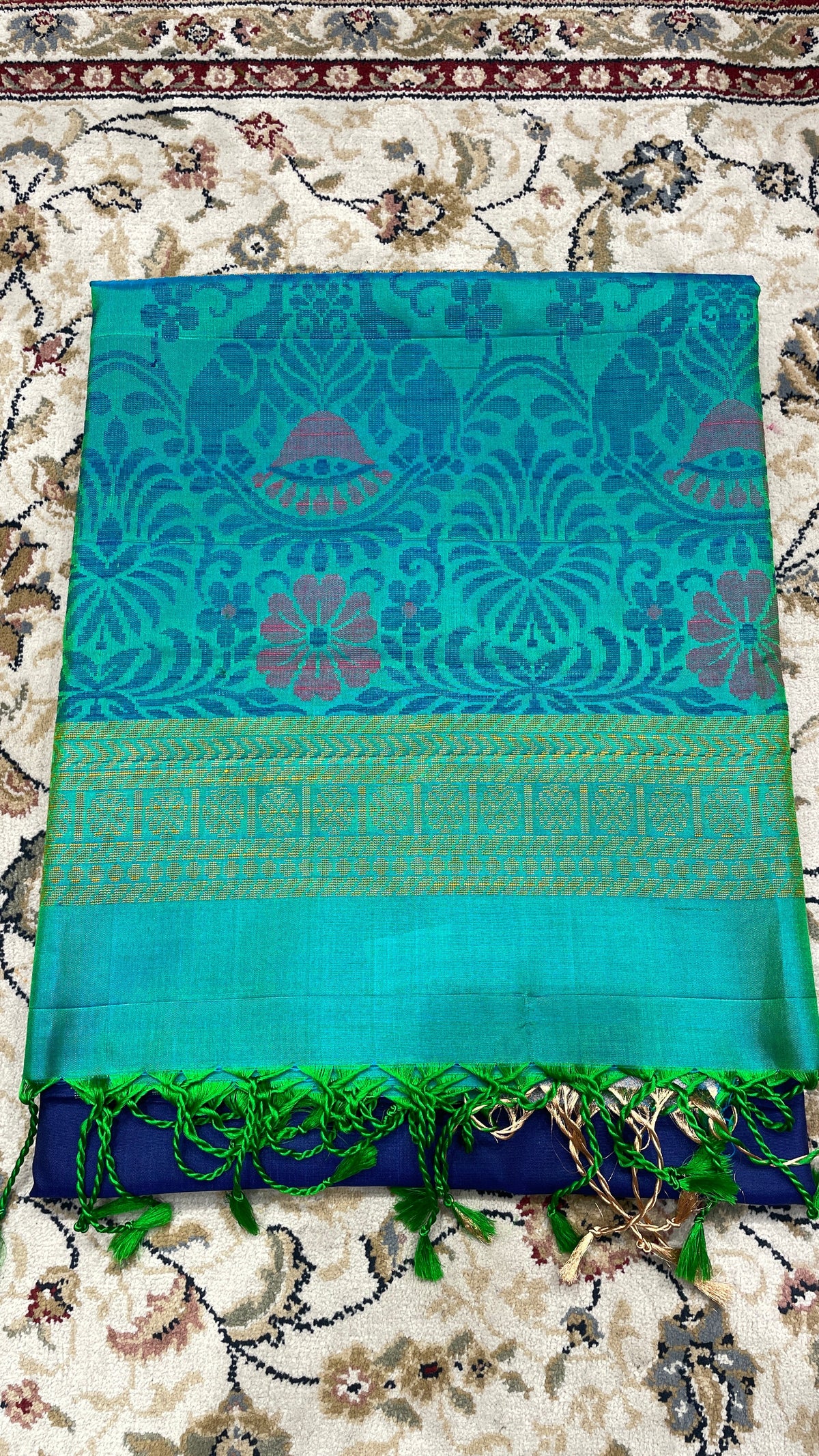 A folded turquoise blue handloom silk saree with intricate floral designs and a green border, displayed on a floral patterned fabric.