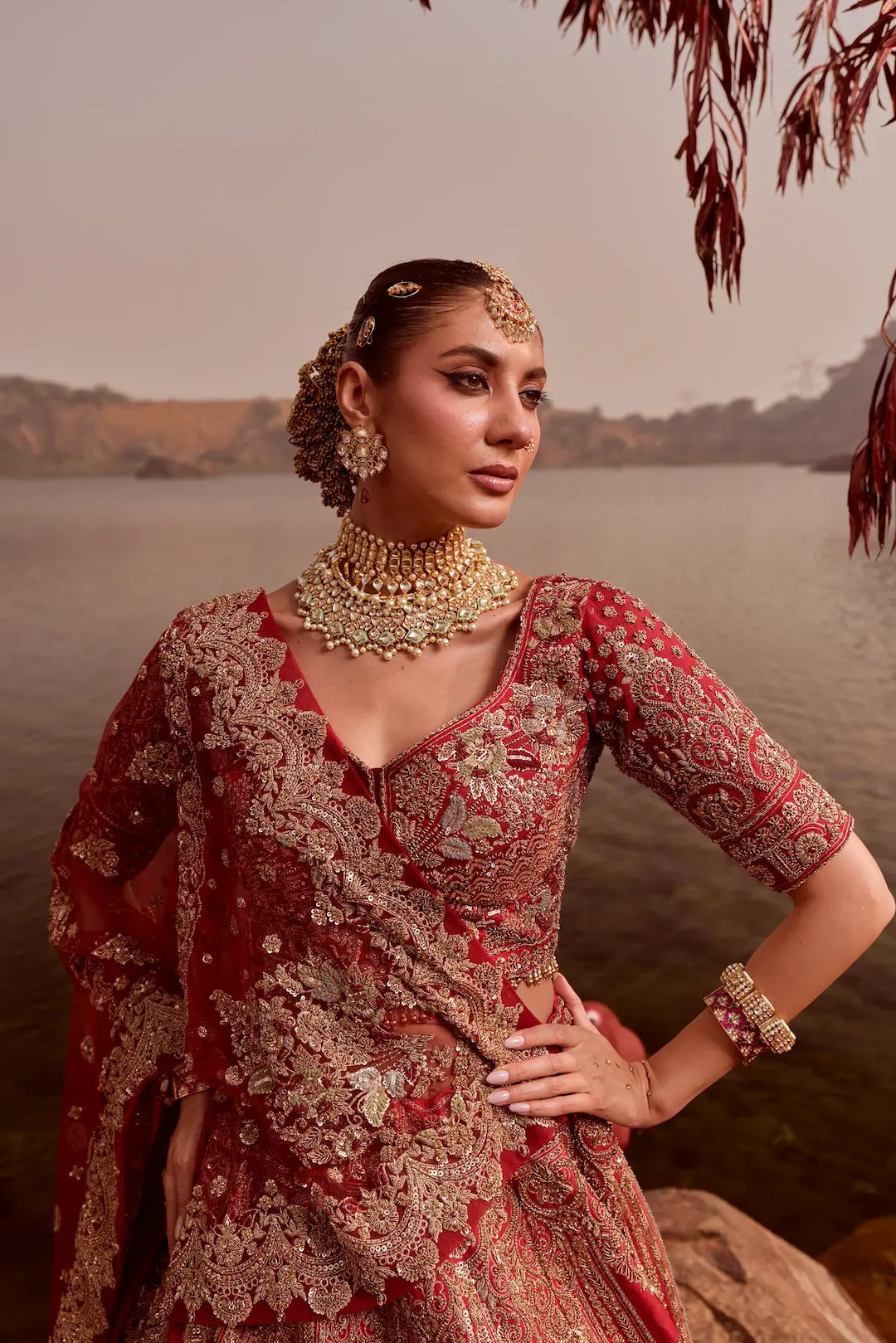 Woman in traditional red and gold outfit with jewelry by a body of water.