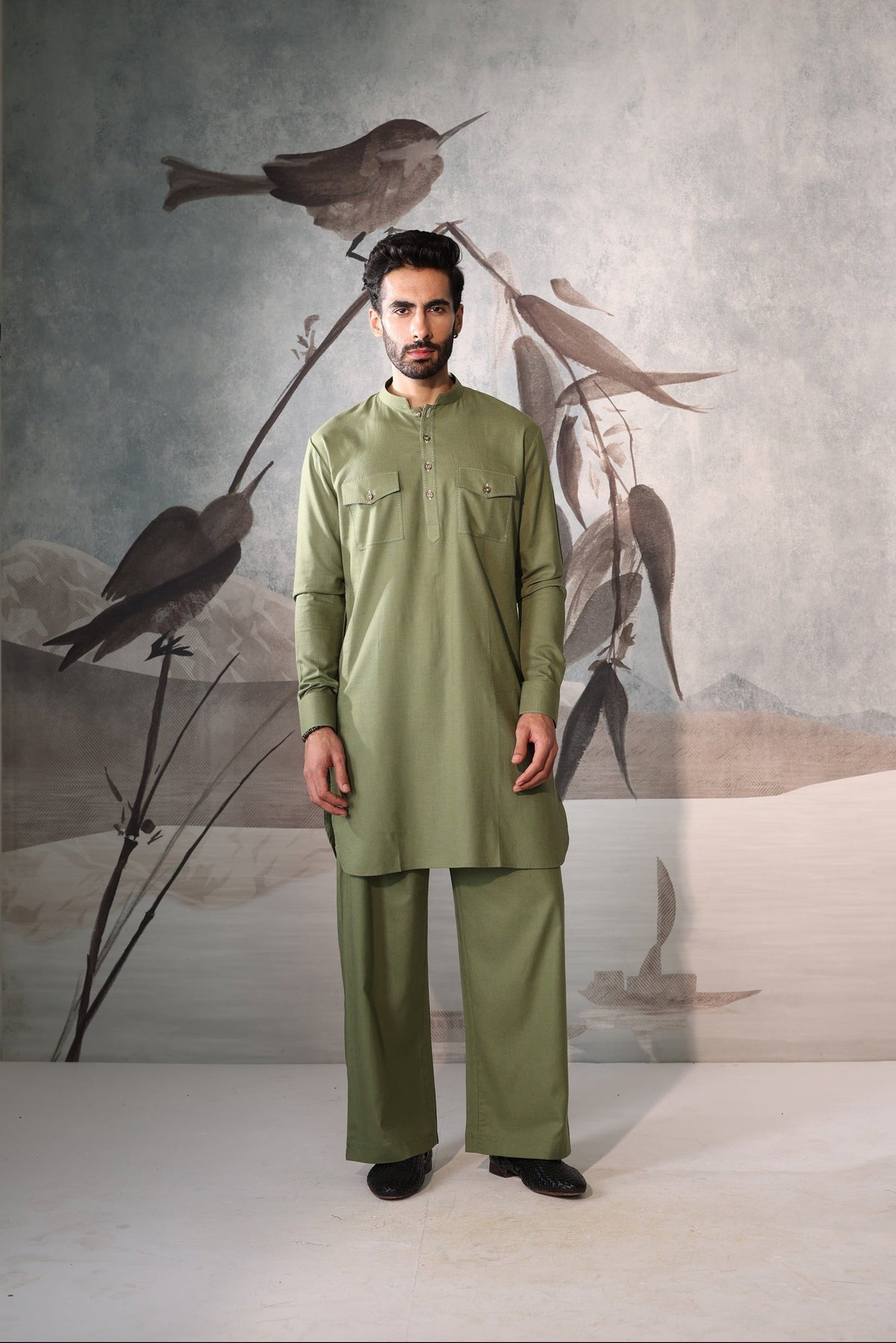 Man wearing a green traditional outfit standing in front of a wall with bird artwork.