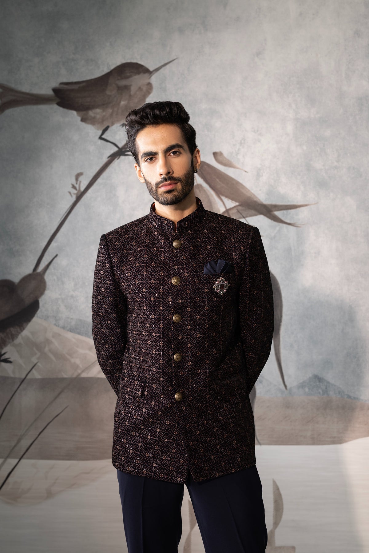 Man wearing a dark patterned jacket in front of a decorative wall with birds.