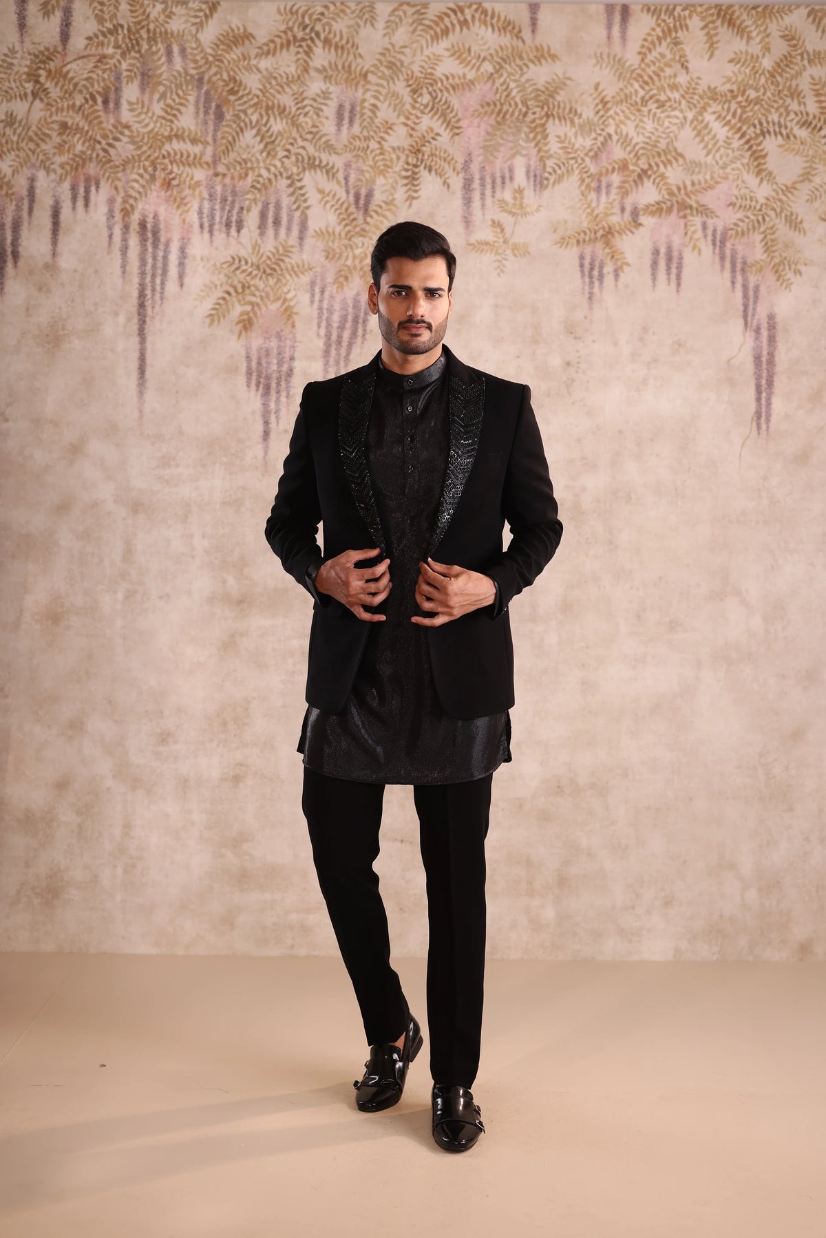 Man in a black suit standing against a decorative wall.