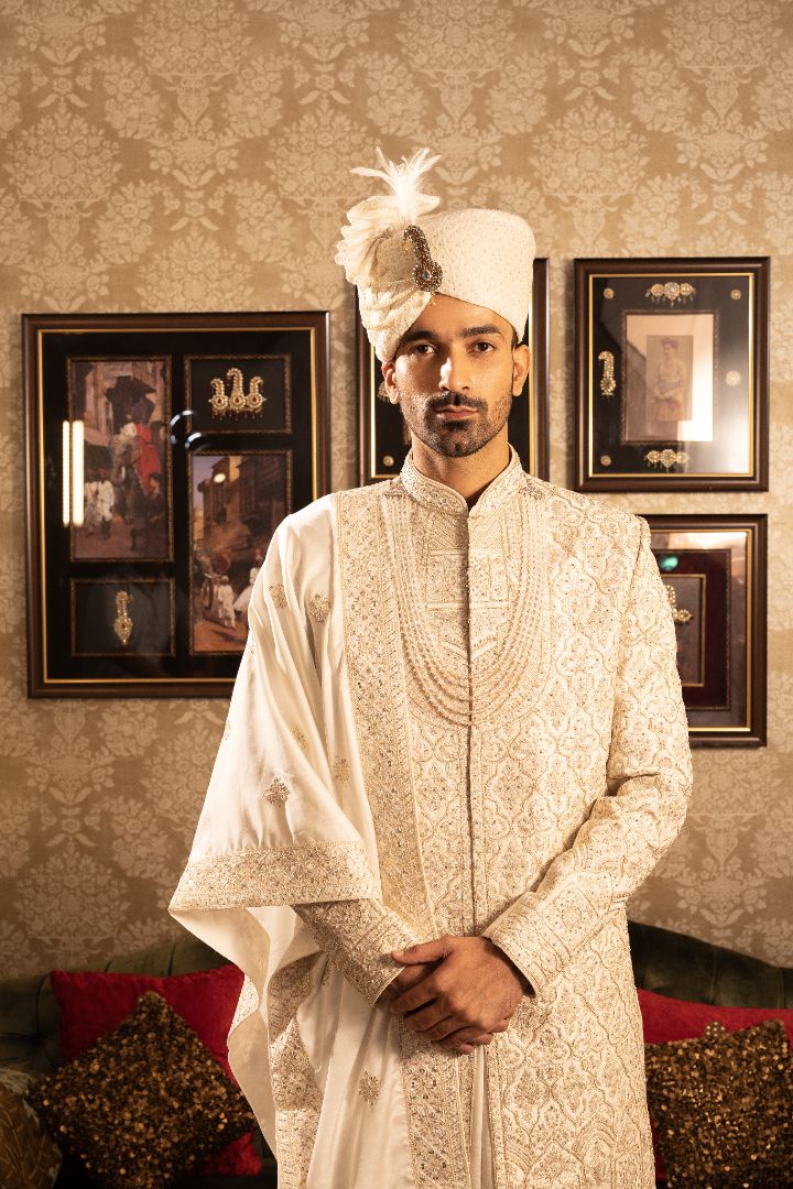 A man wearing a cream-colored sherwani with intricate gold patterns, a white turban, and standing in a room with a rich, ornate background.