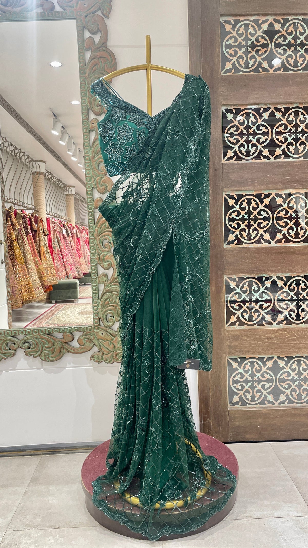 A green net saree with floral embellishments displayed on a mannequin in a store with traditional attire and decorative patterns in the background.