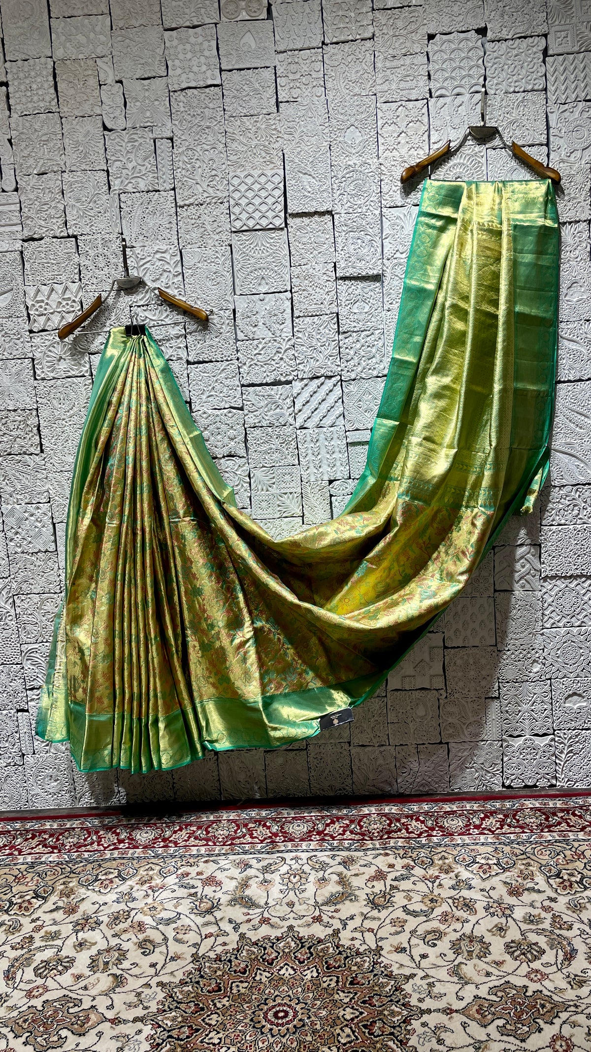 A green silk kanjivaram saree draped over a white textured surface, with part of the saree visible on a wooden hanger against a plain wall.