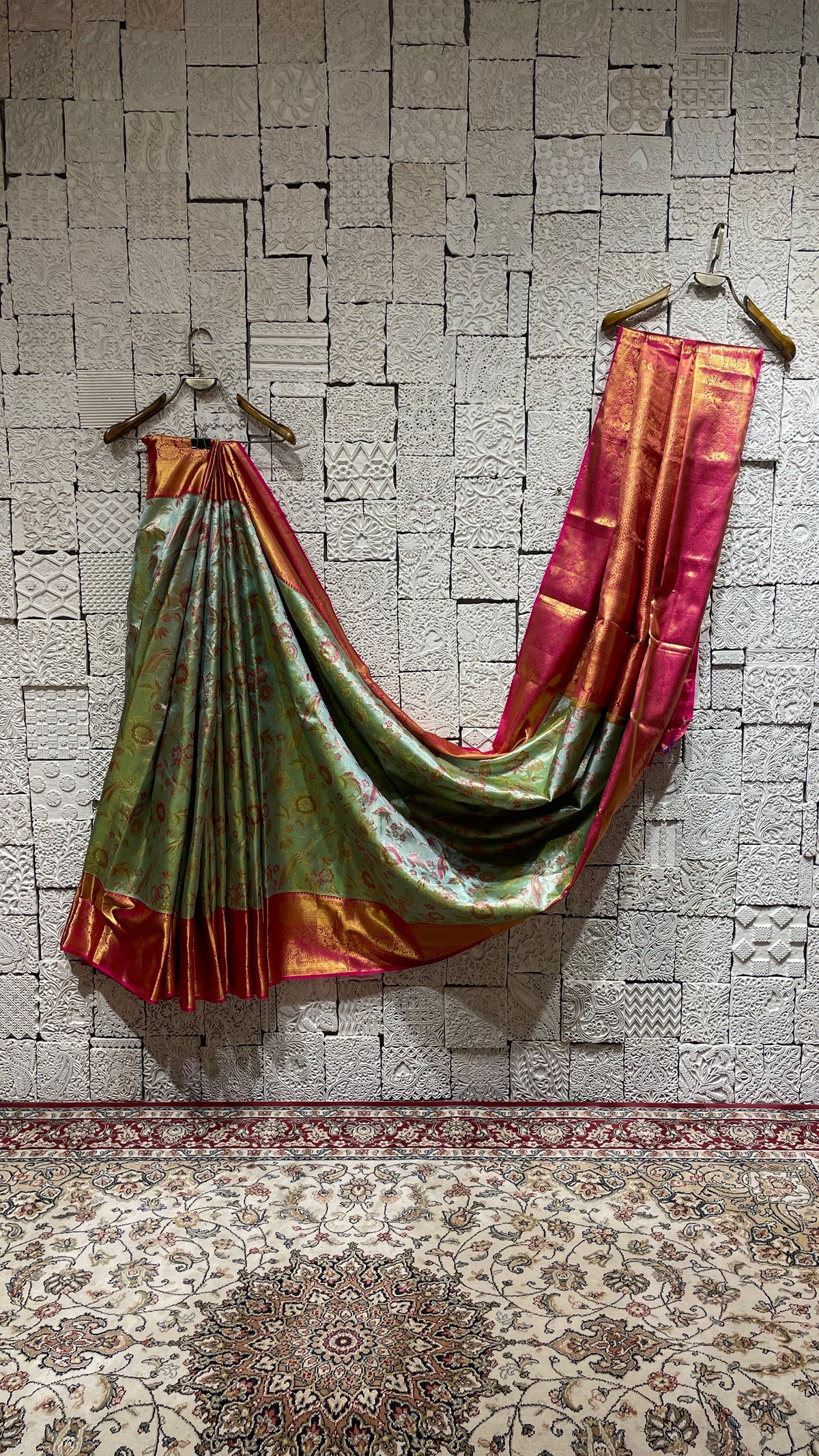 A green silk Kanjivaram saree with a vibrant pink border, draped over a white surface, with a visible zariwork on the pink border.