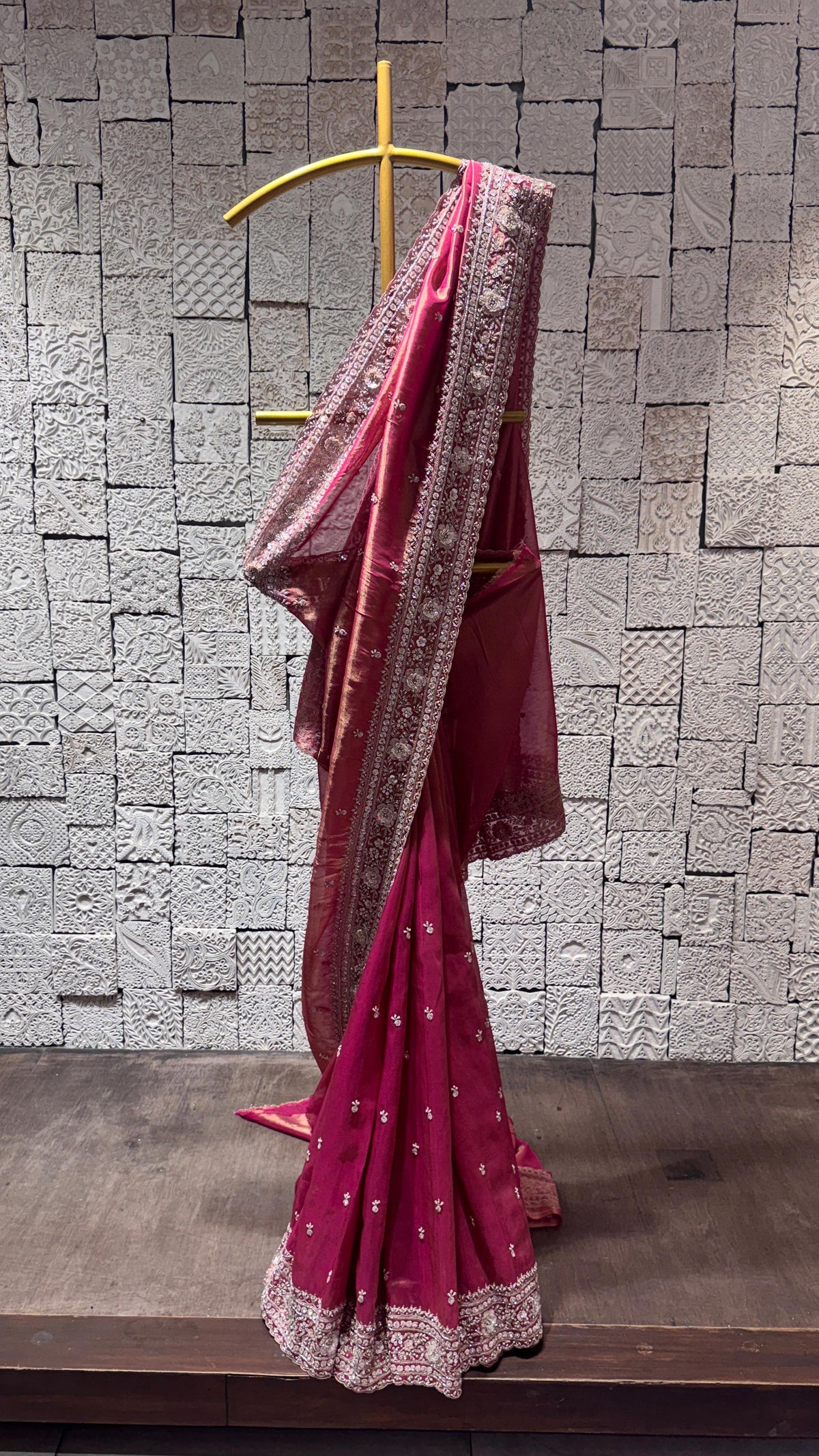A tissue silk saree with heavy sequins border displayed on a hanger against a brick wall background, primarily pink in color with sequin embellishments.